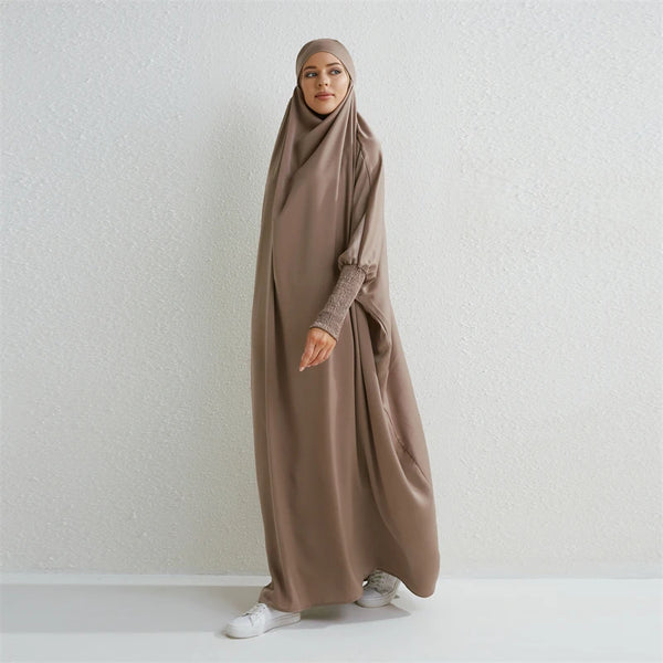 A woman wearing a long, flowing taupe abaya with a smocked sleeve and headscarf.