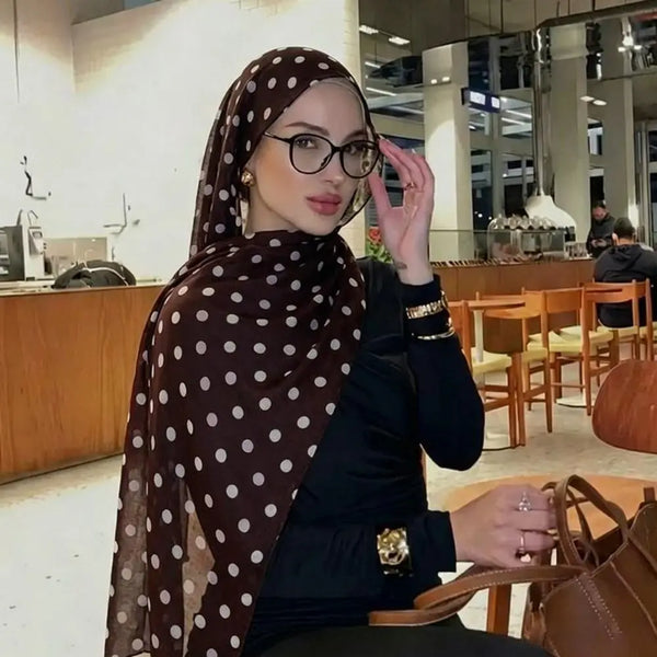 A woman wearing a brown polka dot cotton shawl and glasses sits at a wooden table.