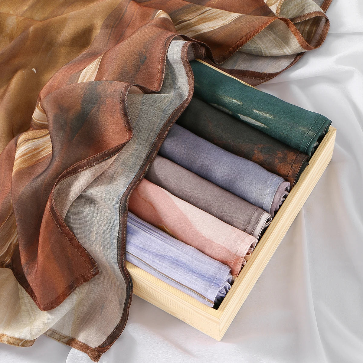A stack of elegant watercolor print modal scarves in various earthy and muted tones, folded inside a light wooden box.