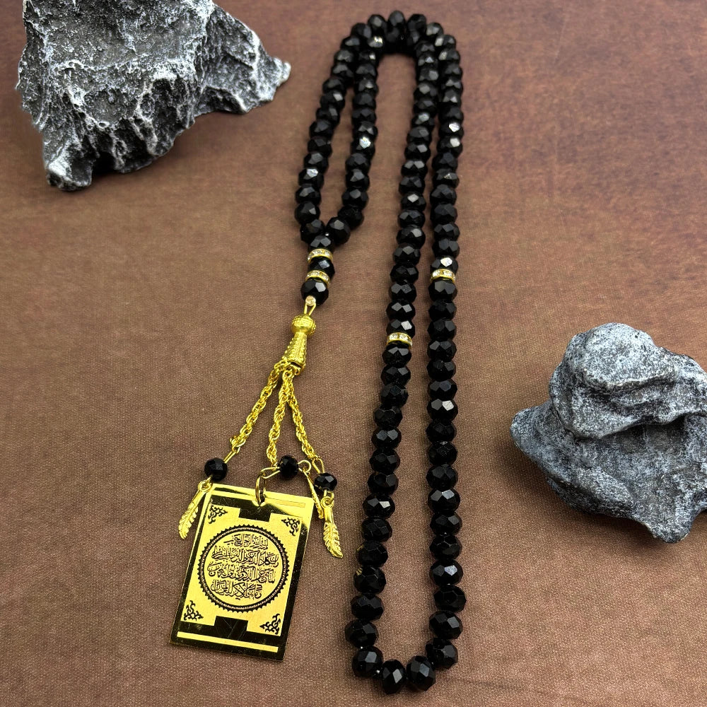 Schwarze Perlenkette mit goldfarbener Plakette auf braunem Stoff neben grauen Steinen.