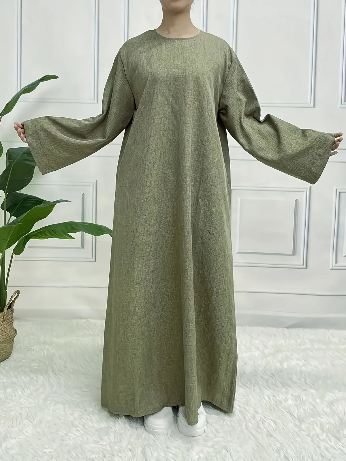 Vestido caftán verde oliva de largo completo con mangas largas y corte holgado, mostrado contra una pared blanca de paneles.