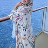 Una mujer con vestido caftán floral y hijab está en un balcón mirando al mar.