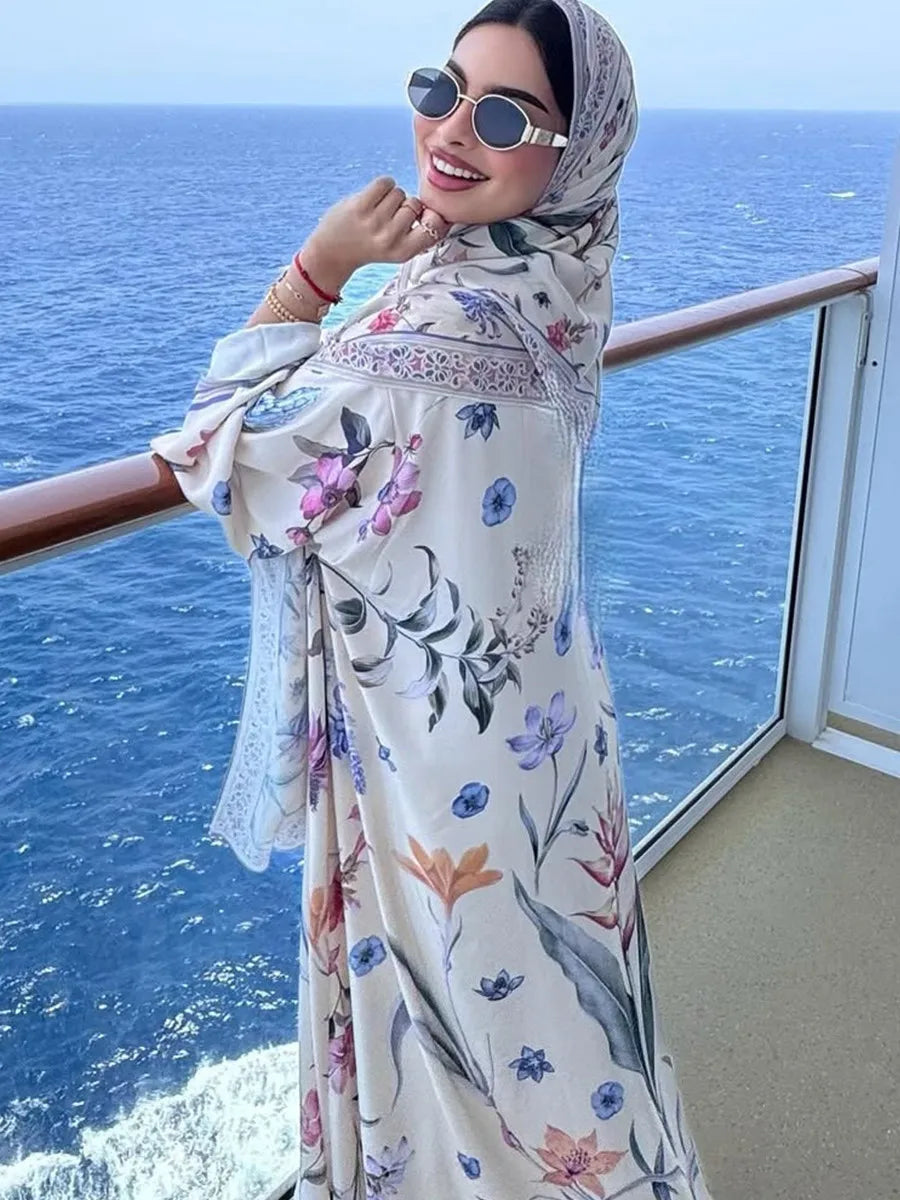 Una mujer con vestido caftán floral y hijab está en un balcón mirando al mar.