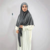 A woman wearing a gray square scarf and white traditional dress, posing against a light wall.