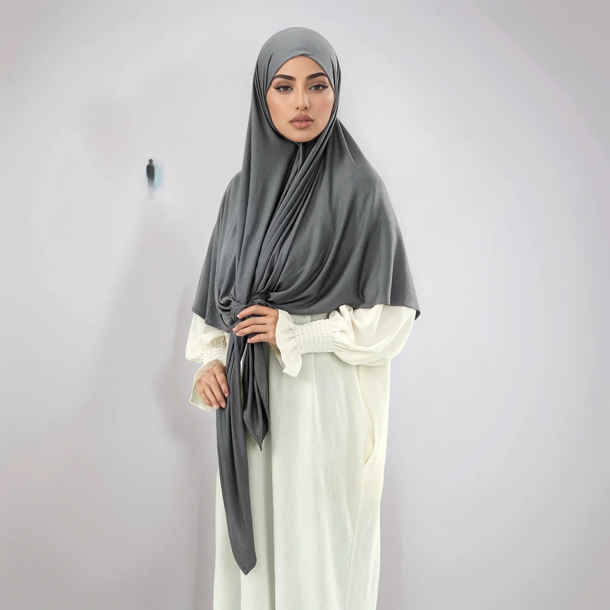 A woman wearing a gray square scarf and white traditional dress, posing against a light wall.