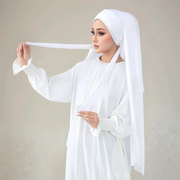 A woman wearing a white instant hijab made of elasticity modal jersey with straps, holding a white fabric strip.