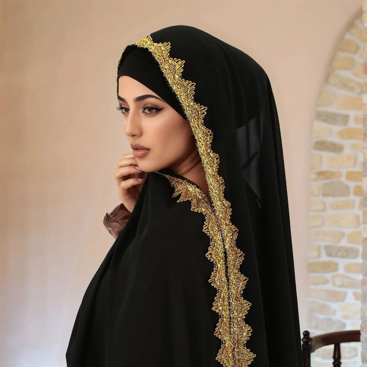 Eine Frau trägt einen schwarzen Spitzen-Chiffon-Hijab mit goldener Stickerei und blickt zur Seite.