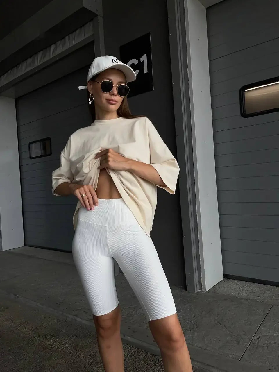 Una mujer viste una camiseta básica de algodón beige oversize y leggings blancos en un pasillo moderno.