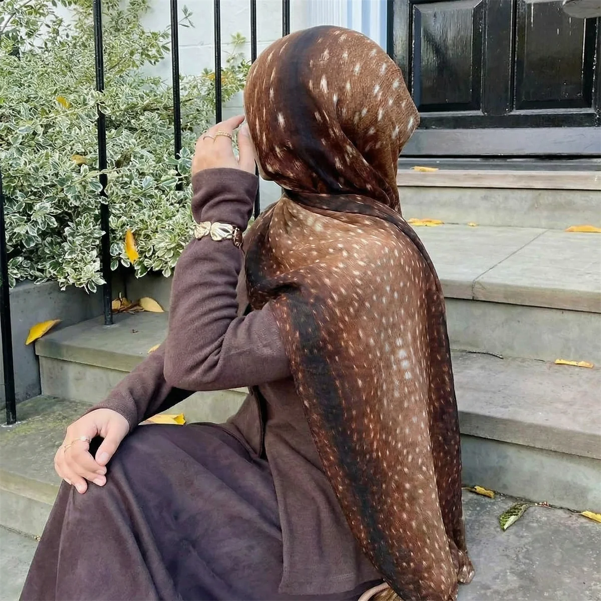 Eine Frau mit braunem Hirschmuster-Hijab-Schal sitzt auf Steinstufen im Freien.