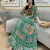 Eine Frau in einem langen, fließenden grünen und orangen Boho-bestickten Maxi-Kleid sitzt auf einem Sofa.
