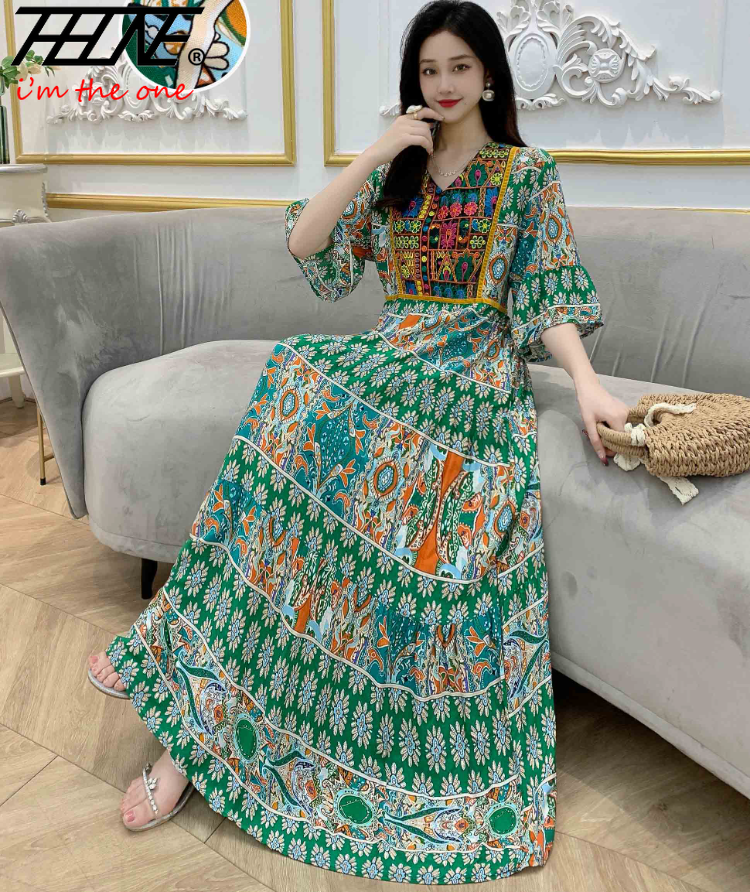 Eine Frau in einem langen, fließenden grünen und orangen Boho-bestickten Maxi-Kleid sitzt auf einem Sofa.