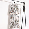 Weißes Maxi-Kleid mit Boho-Blumenmuster an einem schwarzen Haken vor weißer Wand.