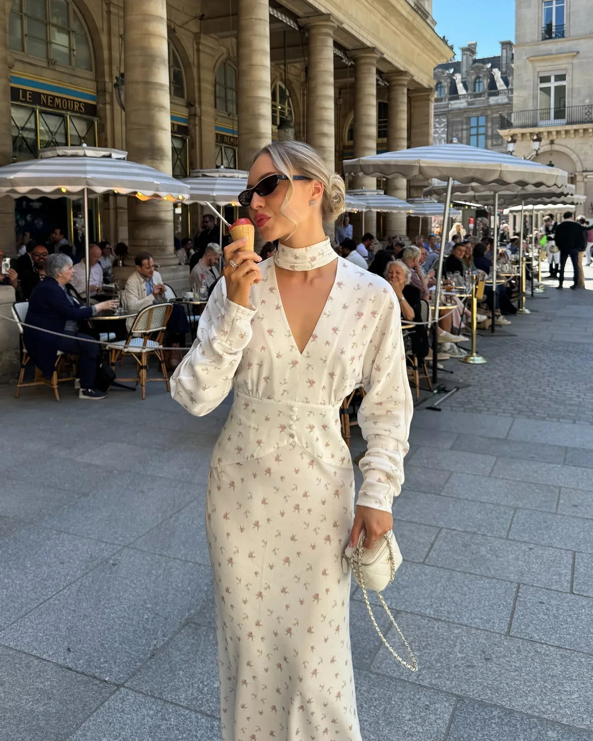 Una mujer vistiendo un vestido largo de satén con estampado floral crema y gafas de sol, parada en una calle peatonal.