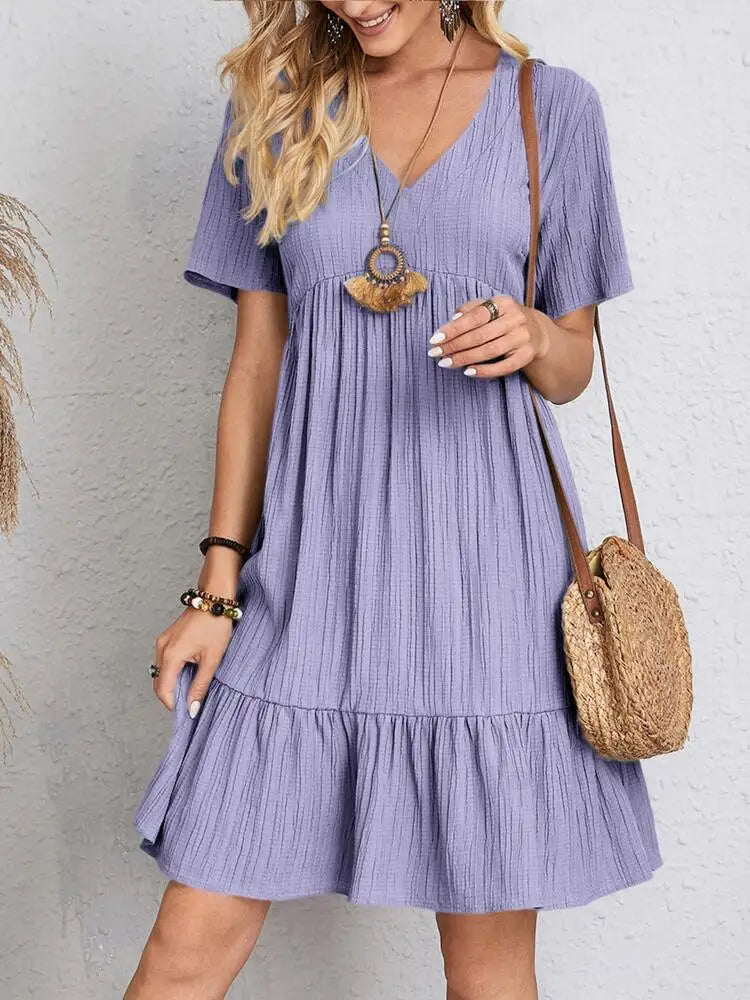 Eine Frau in einem lavendelfarbenen, einfarbigen V-Ausschnitt-Kleid mit A-Linien, gerollten Ärmeln und goldenen Schmuck.