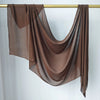 Un foulard hijab en modal marron et transparent drapé, suspendu sur un bâton en bois sur fond blanc.