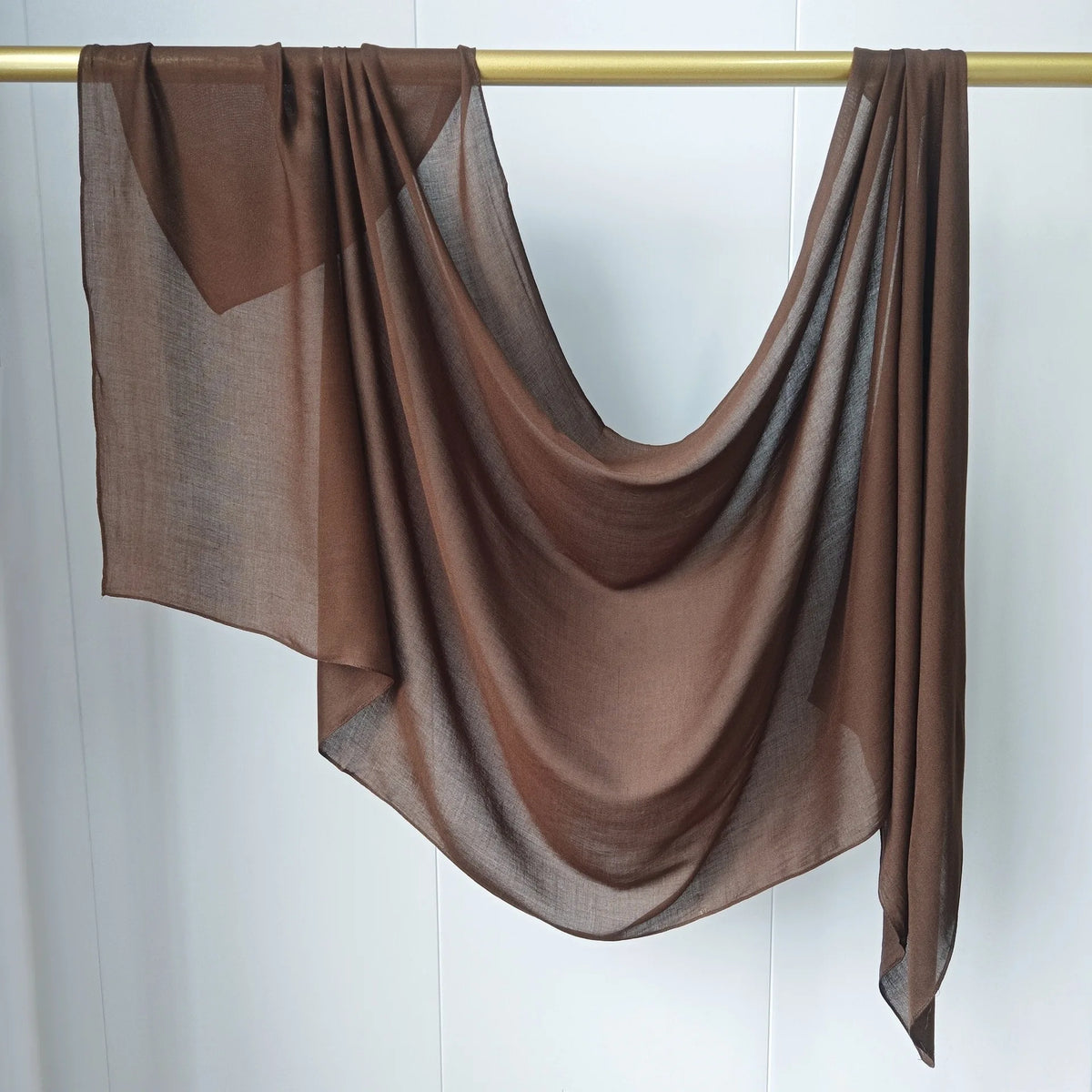Un foulard hijab en modal marron et transparent drapé, suspendu sur un bâton en bois sur fond blanc.