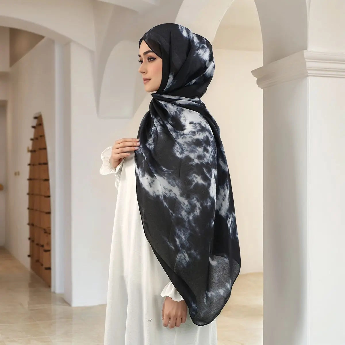 A woman wearing a black and white watercolor print hijab stands in a bright hallway.