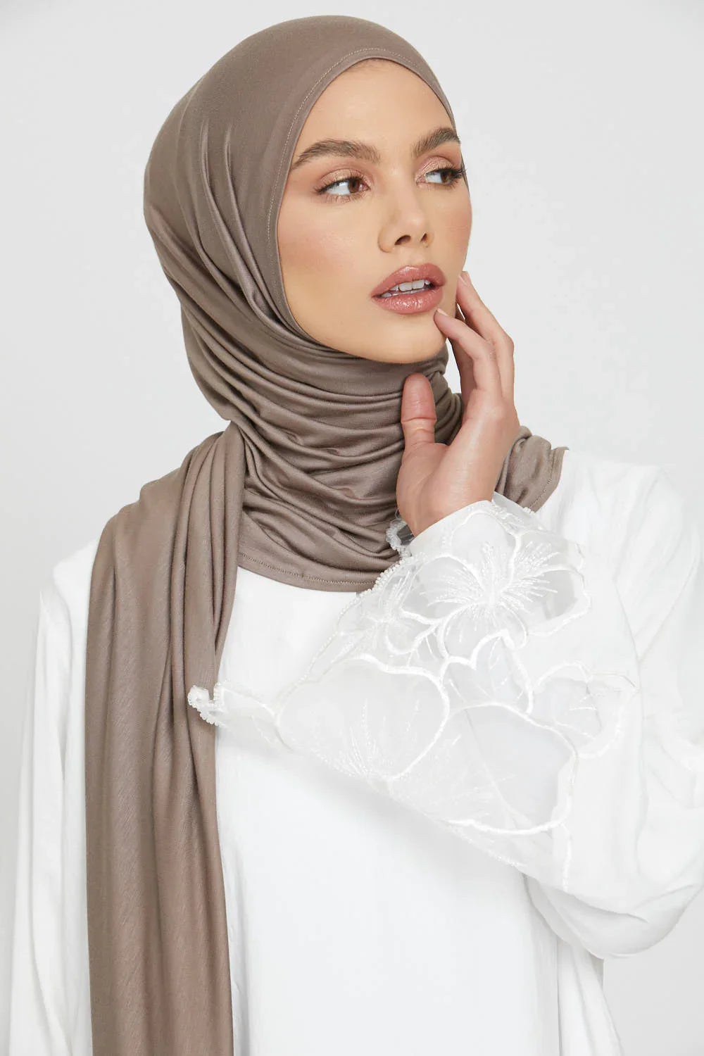 Eine Frau trägt einen einfarbigen braunen Jersey-Hijab und ein weißes Oberteil, blickt zur Seite.