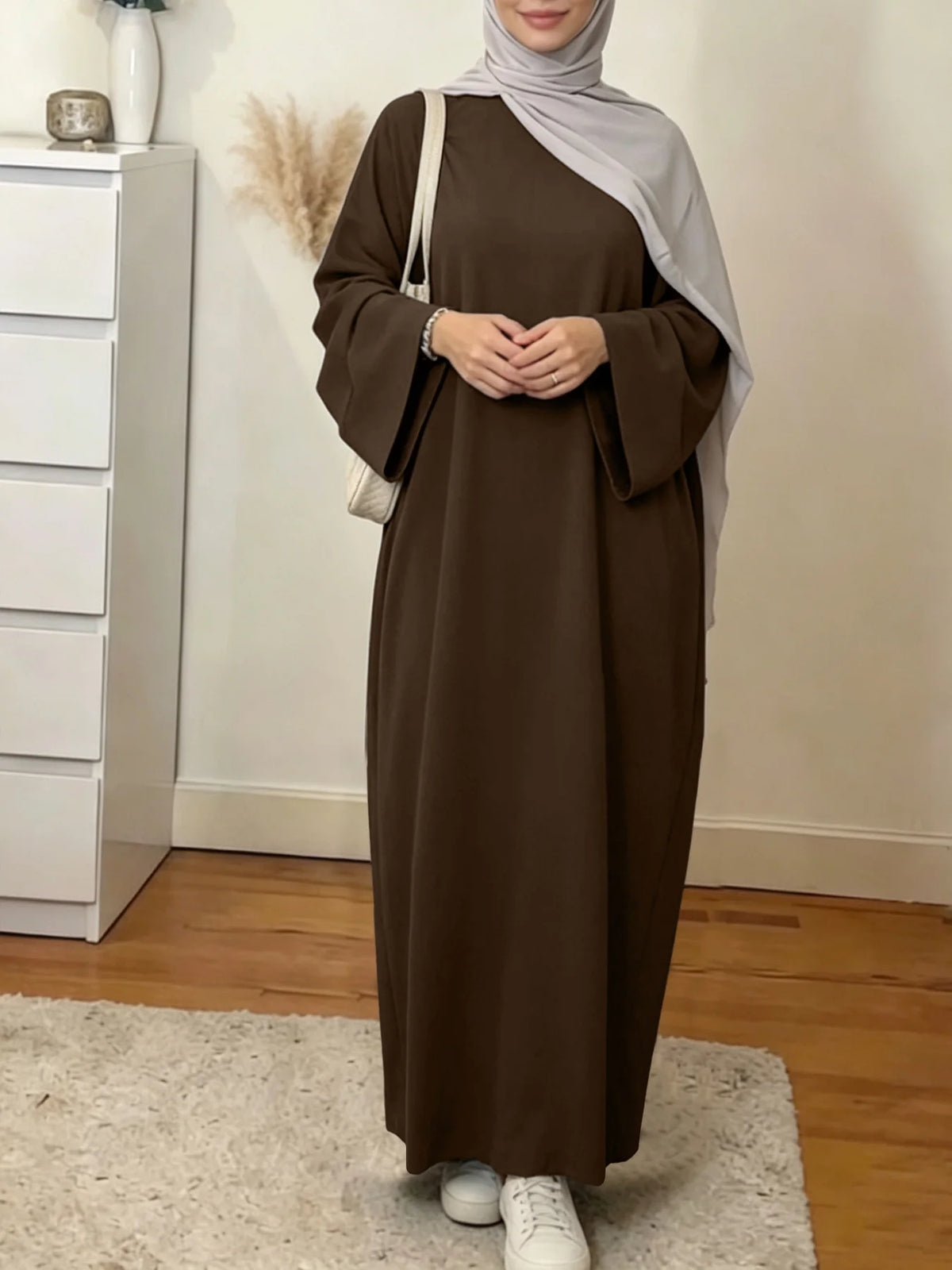 Une femme portant une élégante abaya ample marron et un hijab gris clair, à l'intérieur.