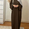 Une femme portant une élégante abaya ample marron uni et un hijab gris clair se tient à l'intérieur.