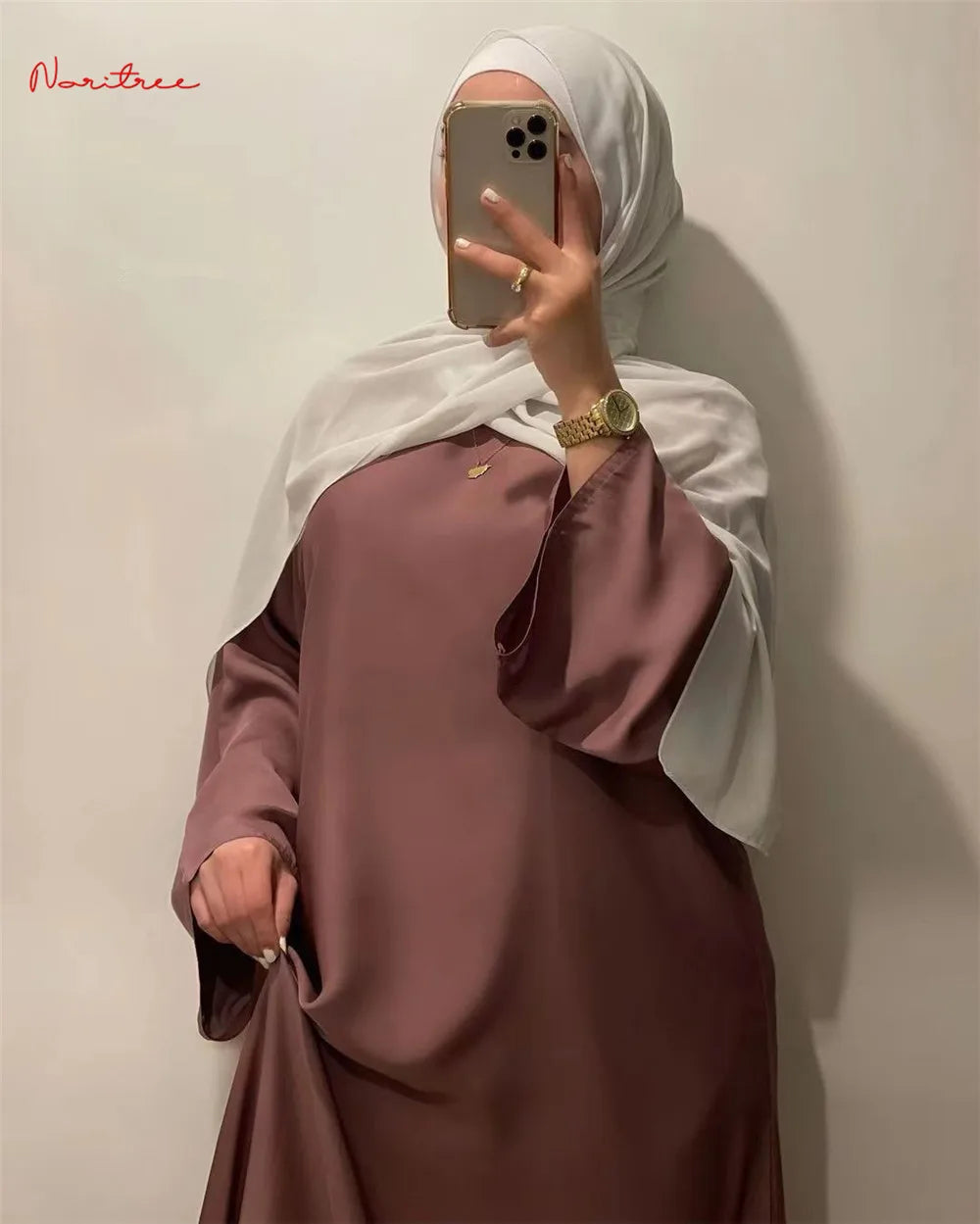 Eine Frau trägt eine mauvefarbene, fließende, lange Abaya mit weiten Ärmeln und einen weißen Hijab und macht ein Spiegelselfi