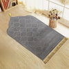Tapis de prière gris embossé avec une pompon doré posé sur un plancher en bois clair.