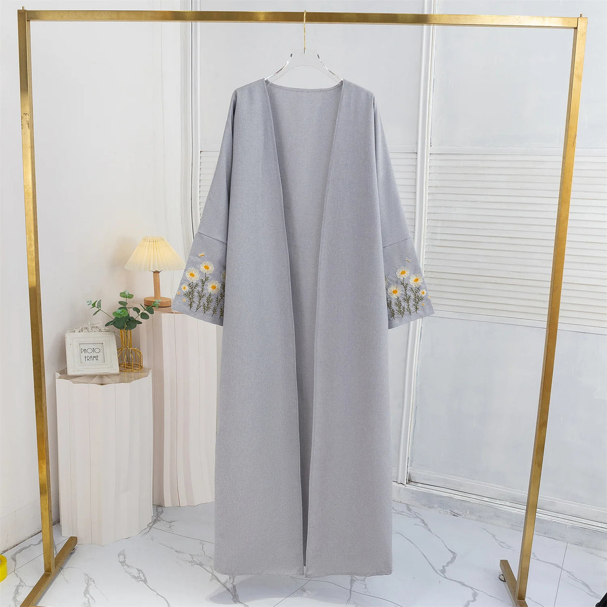 Lange, graue offene Abaya mit floraler Stickerei auf einem goldenen Haken in einem hellen Raum.