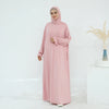Una mujer vistiendo un vestido rosa largo y fluido y hijab, de pie contra un fondo blanco.