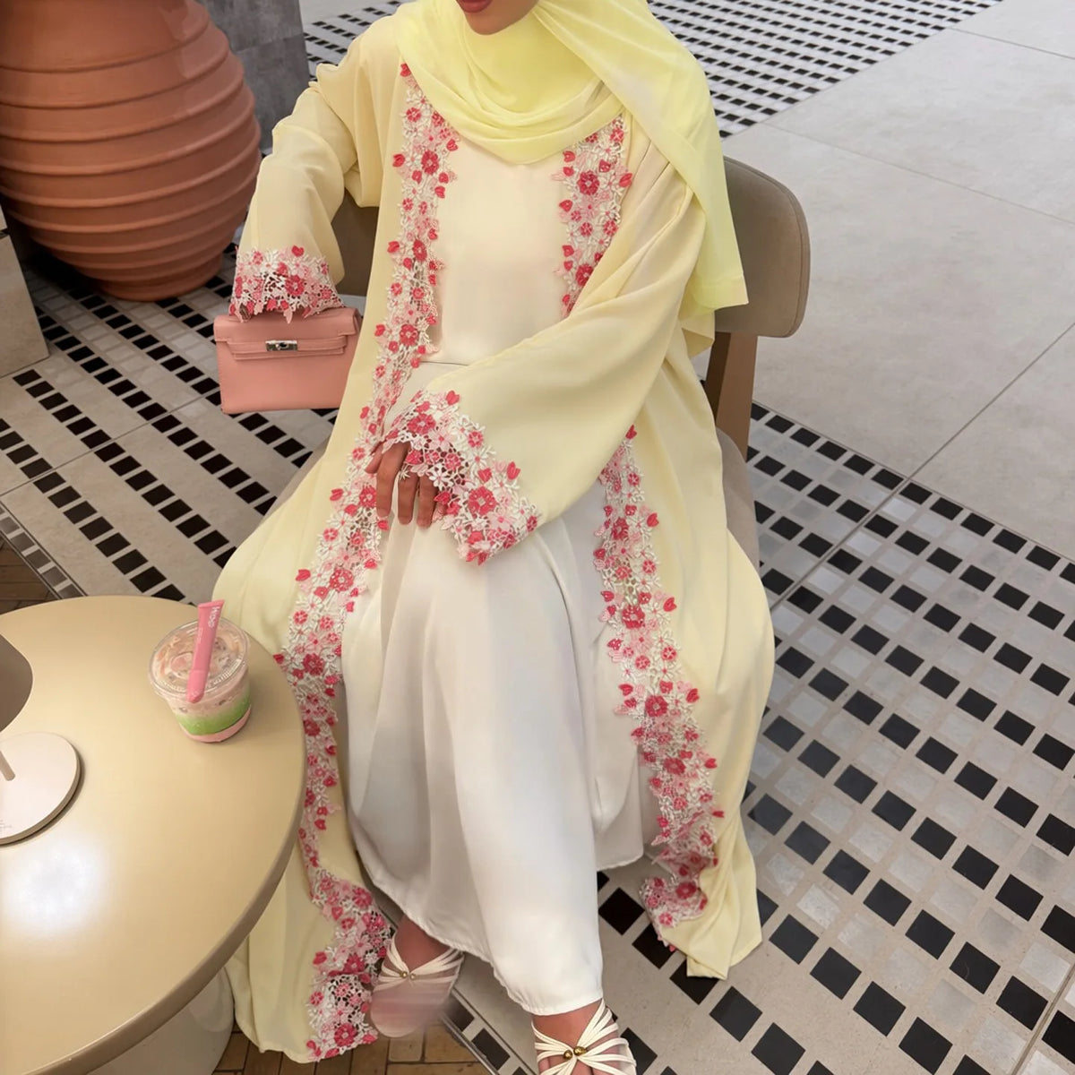 Une femme portant une abaya crème avec broderie de dentelle florale et un hijab jaune, debout à l'intérieur.