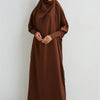 Eine Frau trägt eine braune, bescheidene Abaya mit gerafften Ärmeln und einem passenden Hijab vor hellem Hintergrund.