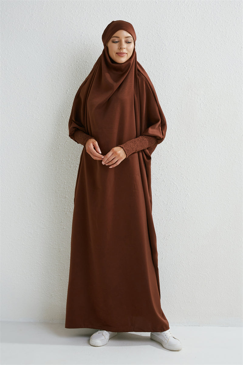 Eine Frau trägt eine braune, bescheidene Abaya mit gerafften Ärmeln und einem passenden Hijab vor hellem Hintergrund.
