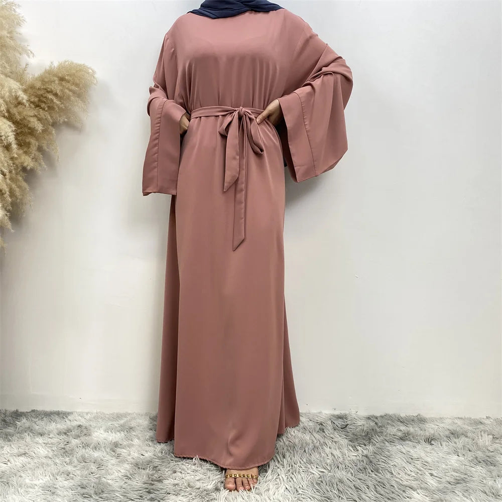 Abaya de color rosa empolvado para mujer, de manga larga y cintura atada, mostrada contra una pared blanca.