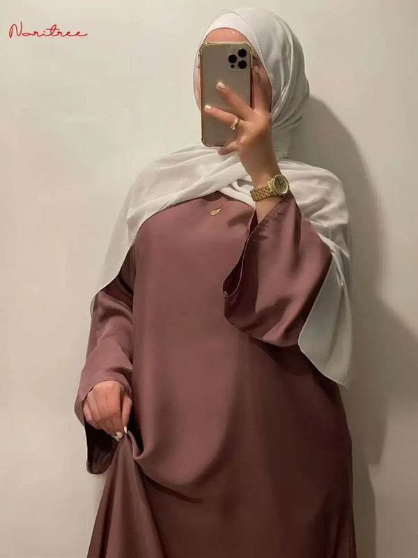A woman wearing a mauve flowy full-length abaya with flare sleeves and a white hijab, posing for a photo.