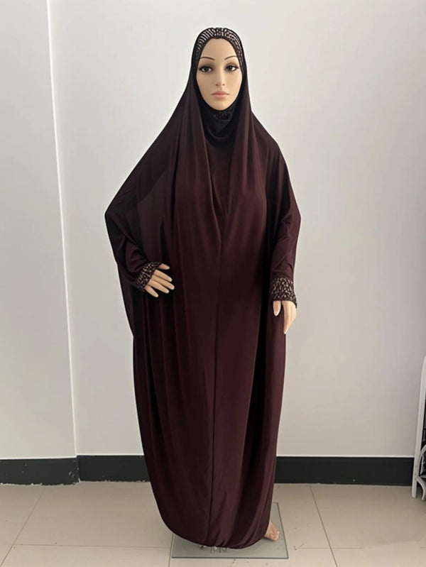 A model wearing a dark maroon flowy kaftan abaya dress with a black hijab and patterned cuffs.