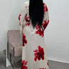 A woman wearing a long, flowing ivory kaftan dress with a vibrant red floral print, standing against a white wall.