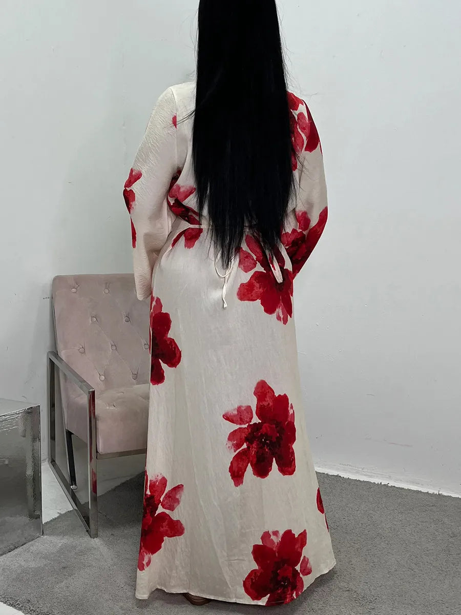 A woman wearing a long, flowing ivory kaftan dress with a vibrant red floral print, standing against a white wall.