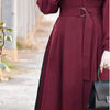 Una mujer vistiendo un vestido maxi holgado color granate de manga larga y cintura ceñida, sosteniendo un bolso negro.