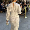 Una mujer vistiendo un vestido largo de satén estampado floral crema con cuello alto y espalda abierta, de pie al aire libre.