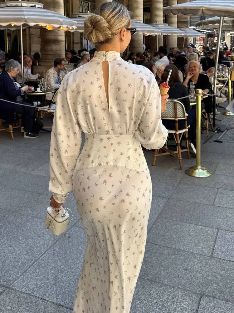 Una mujer vistiendo un vestido largo de satén estampado floral crema con cuello alto y espalda abierta, de pie al aire libre.
