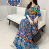 Eine Frau in einem blau-orangefarbenen, bohemieninspirierten, bestickten Maxi-Kleid sitzt auf einem weißen Sofa.