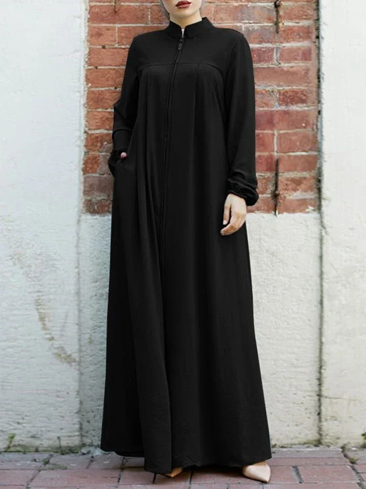 Une femme portant une élégante robe longue noire ample avec col montant, debout contre un mur de briques.