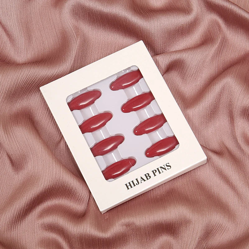 Una caja blanca con ocho pines y clips rojos para hijab, sobre tela de satén rosa.