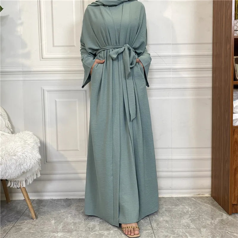 A long, flowing sage green abaya or kaftan set worn by a model in a light-colored room.
