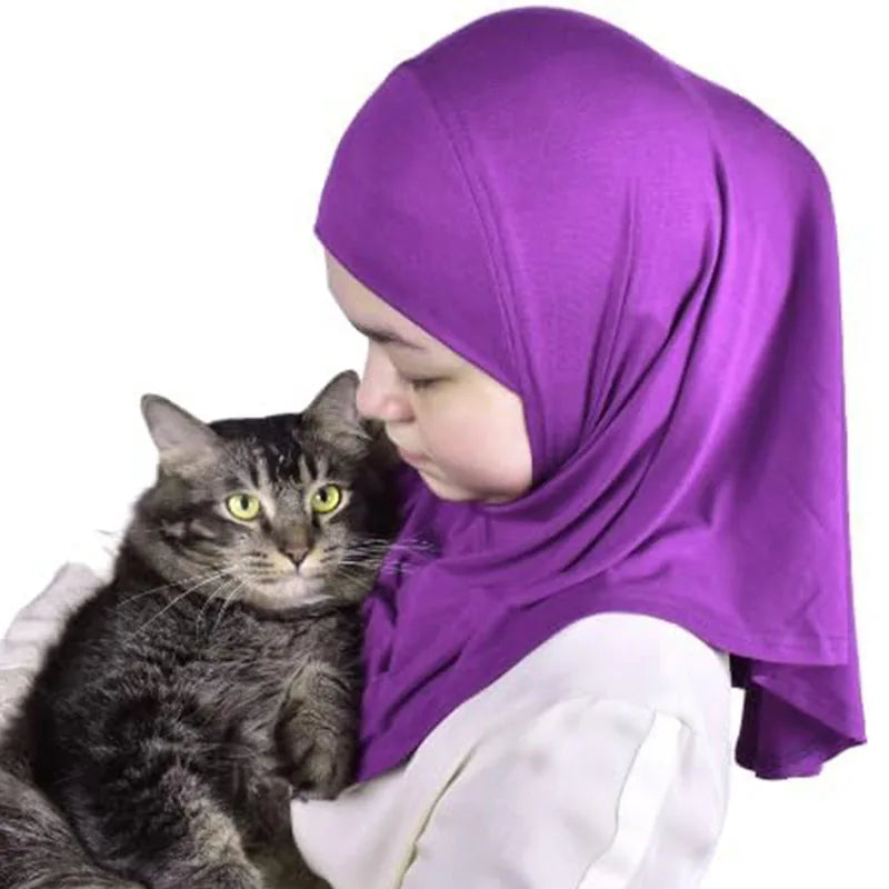 Una niña vistiendo un pañuelo instantáneo musulmán morado mientras sostiene un gatito atigrado.
