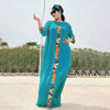 Eine Frau in einem türkisen Kaftan-Set mit farbenfroher Stickerei steht draußen vor einer trockenen Landschaft.