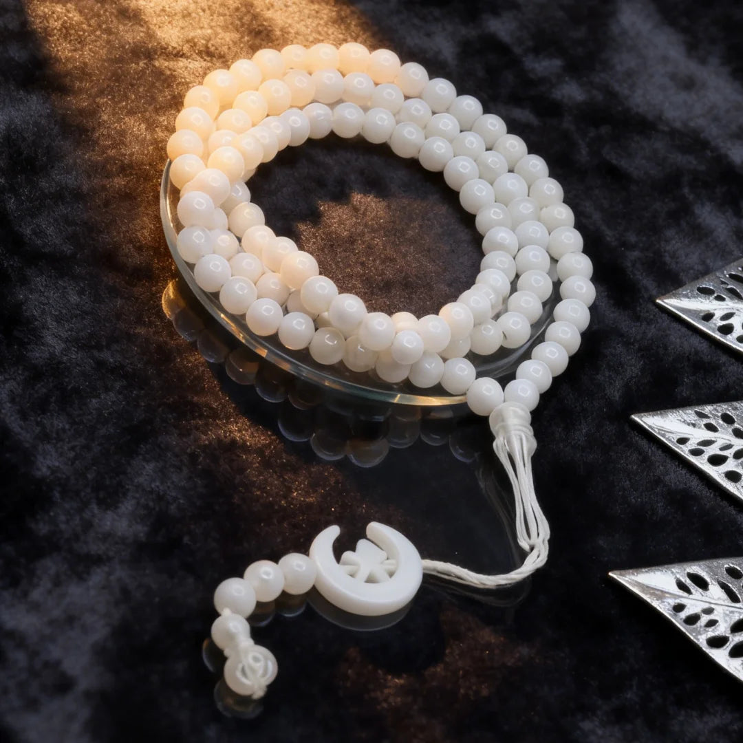 Tasbih de cuentas blanco con borlas de luna y estrella sobre una superficie oscura.