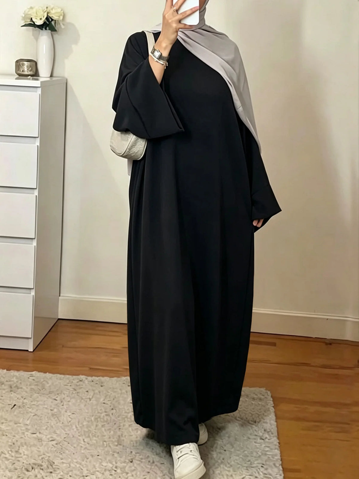 Une femme portant une élégante abaya noire ample et fluide avec un hijab gris et des accents argentés.