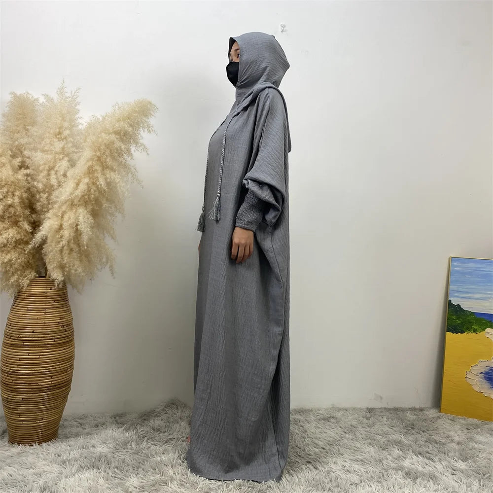Une abaya grise à toute longueur avec capuche et manches batwing, exposée contre un mur blanc.