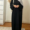 Une femme portant une élégante abaya noire ample et fluide et un hijab gris clair, à l'intérieur.