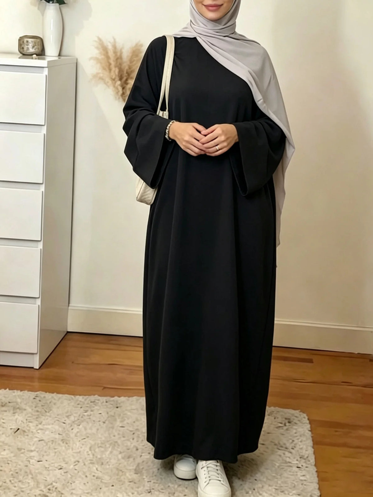 Une femme portant une élégante abaya noire ample et fluide et un hijab gris clair, à l'intérieur.